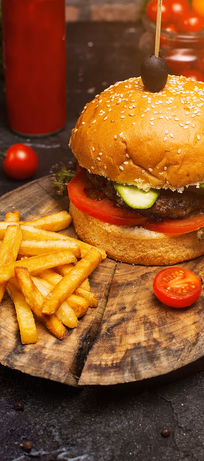 Close-up of a sesame seed hamburger with tomato, cucumber, and a black olive on top, served with French fries on a wooden board.